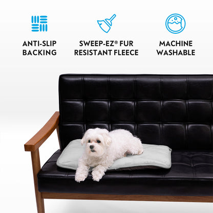 A small white dog on a grey Paw Inspired self-heating pet mat placed on a black leather couch, featuring anti-slip backing, Sweep-EZ fleece, and machine washable icons.