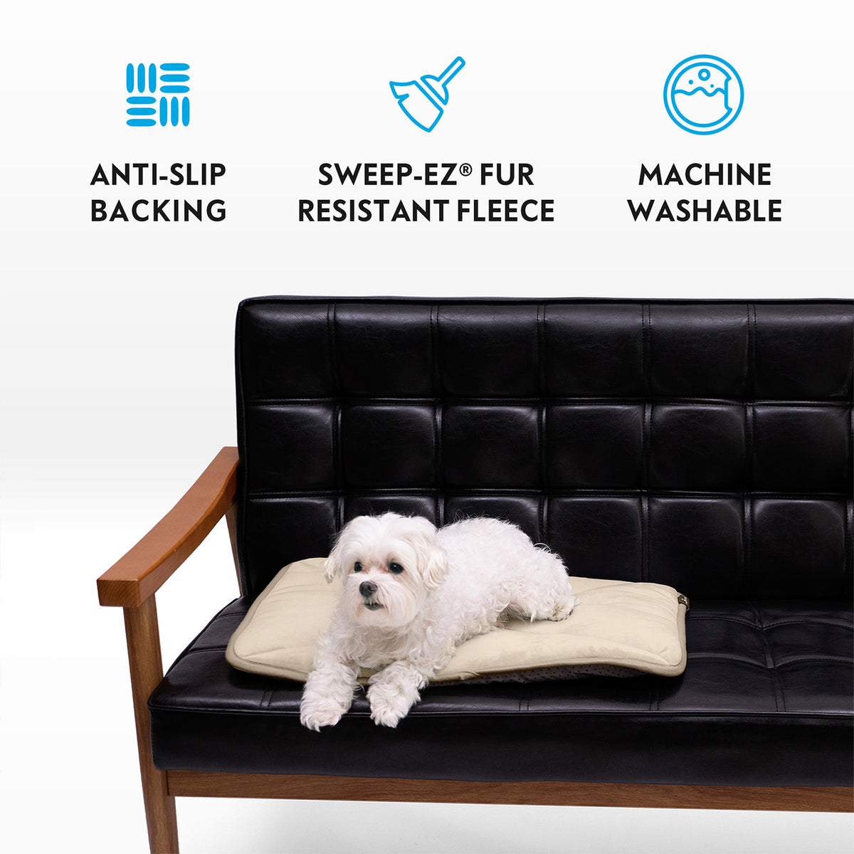 A small white dog on a tan Paw Inspired self-heating pet mat placed on a black leather couch, featuring anti-slip backing, Sweep-EZ fleece, and machine washable icons.