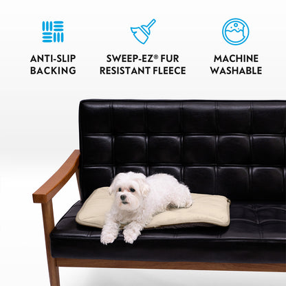 A small white dog on a tan Paw Inspired self-heating pet mat placed on a black leather couch, featuring anti-slip backing, Sweep-EZ fleece, and machine washable icons.