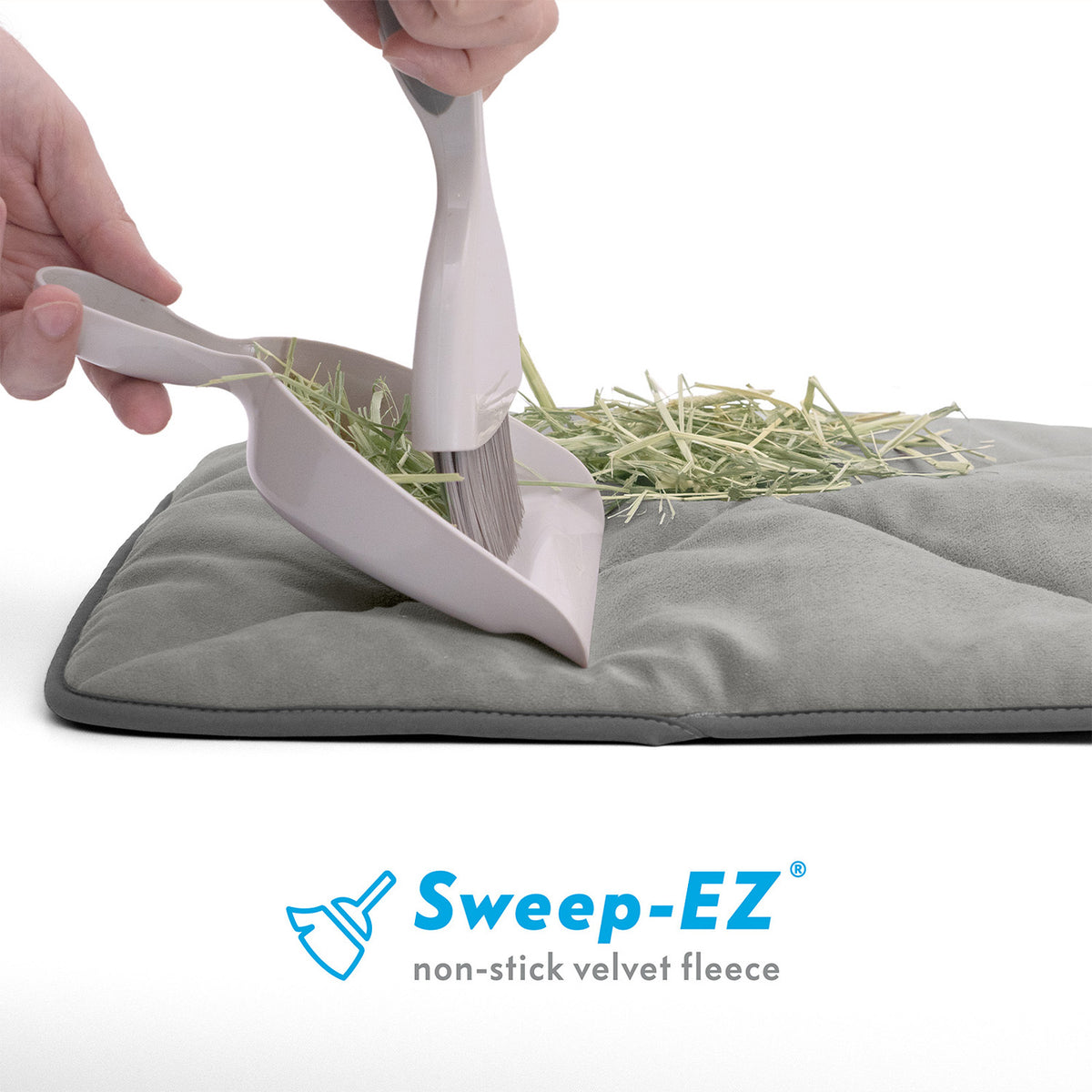 A hand sweeping hay off a grey Paw Inspired self-heating pet mat using a dustpan and brush to show the Sweep-EZ non-stick velvet fleece surface.